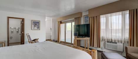 A hotel room with a large bed, a television on a wooden stand, a chair, and an air conditioning unit.