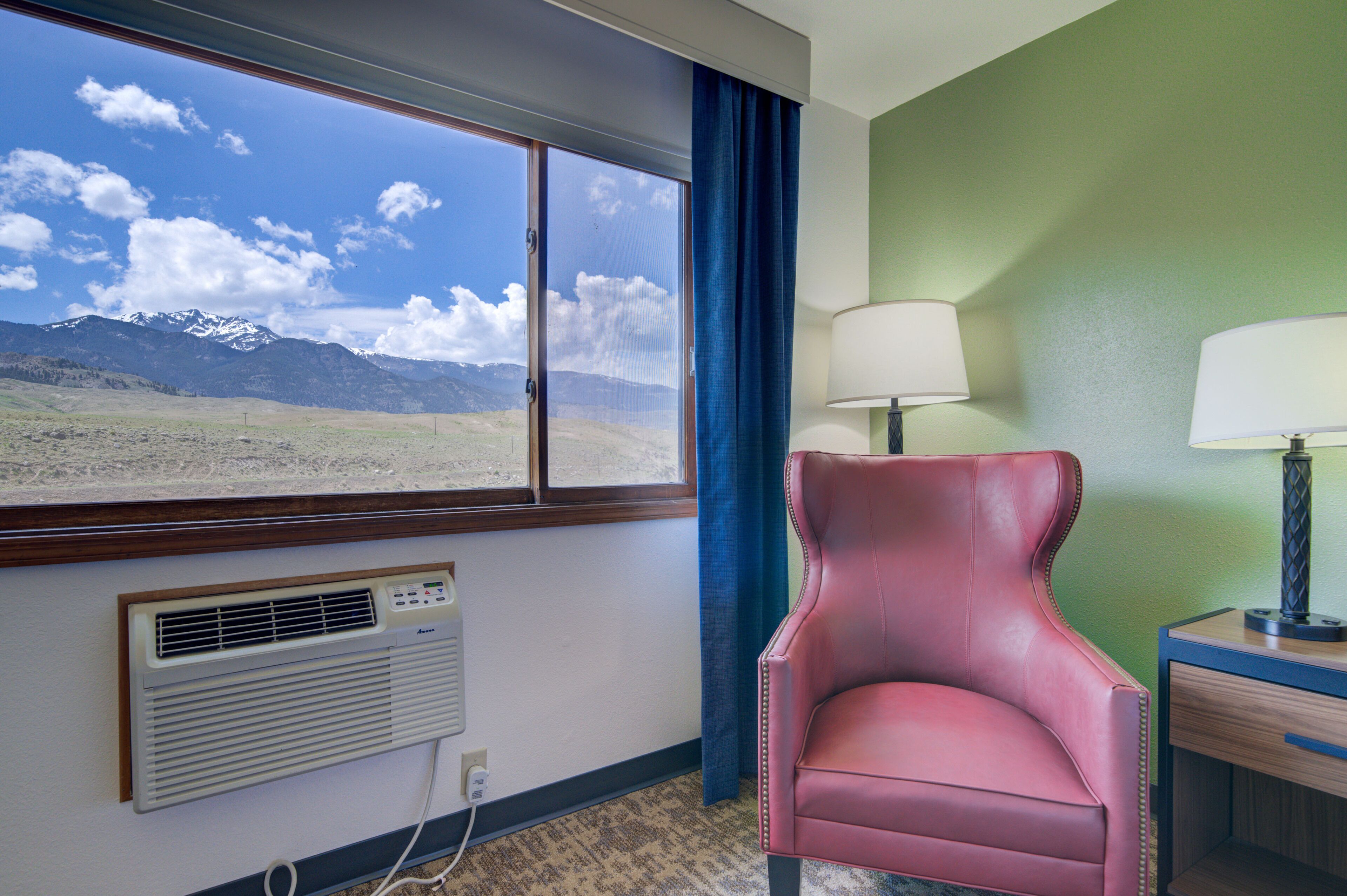 A hotel room with a large window offering a mountain view, a red leather chair, a lamp, and a bedside table.