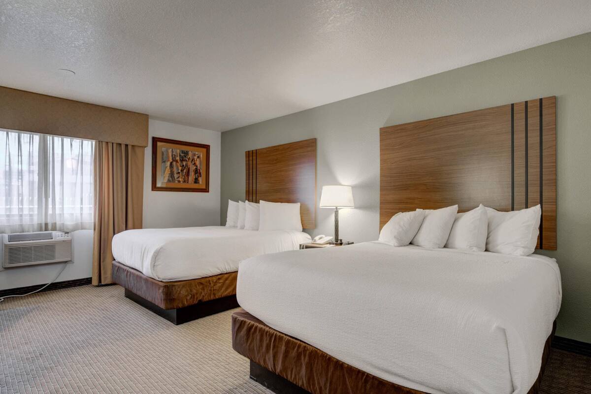 A hotel room with two beds, a large window with curtains, an air conditioning unit, and a painting on the wall.