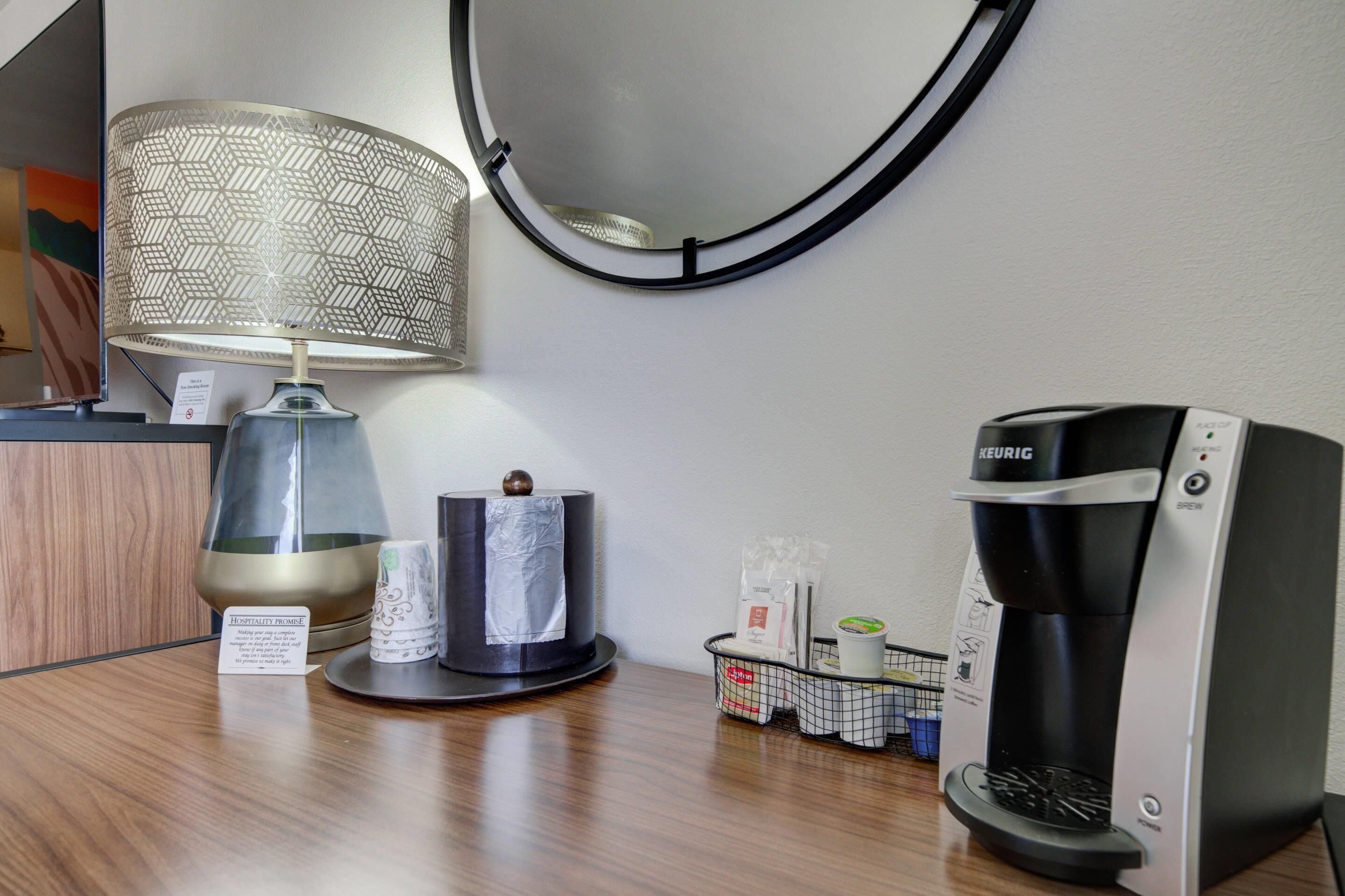 A hotel room with a round mirror, a table lamp, a Keurig coffee maker, and a tissue dispenser.