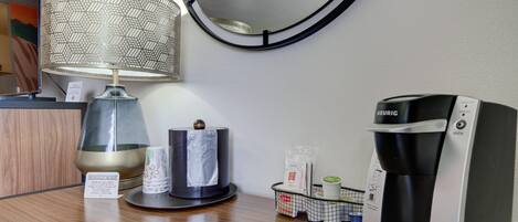 Ein Hotelzimmer mit rundem Spiegel, einer Tischlampe, einer Keurig-Kaffeemaschine und einem Taschentuchspender.