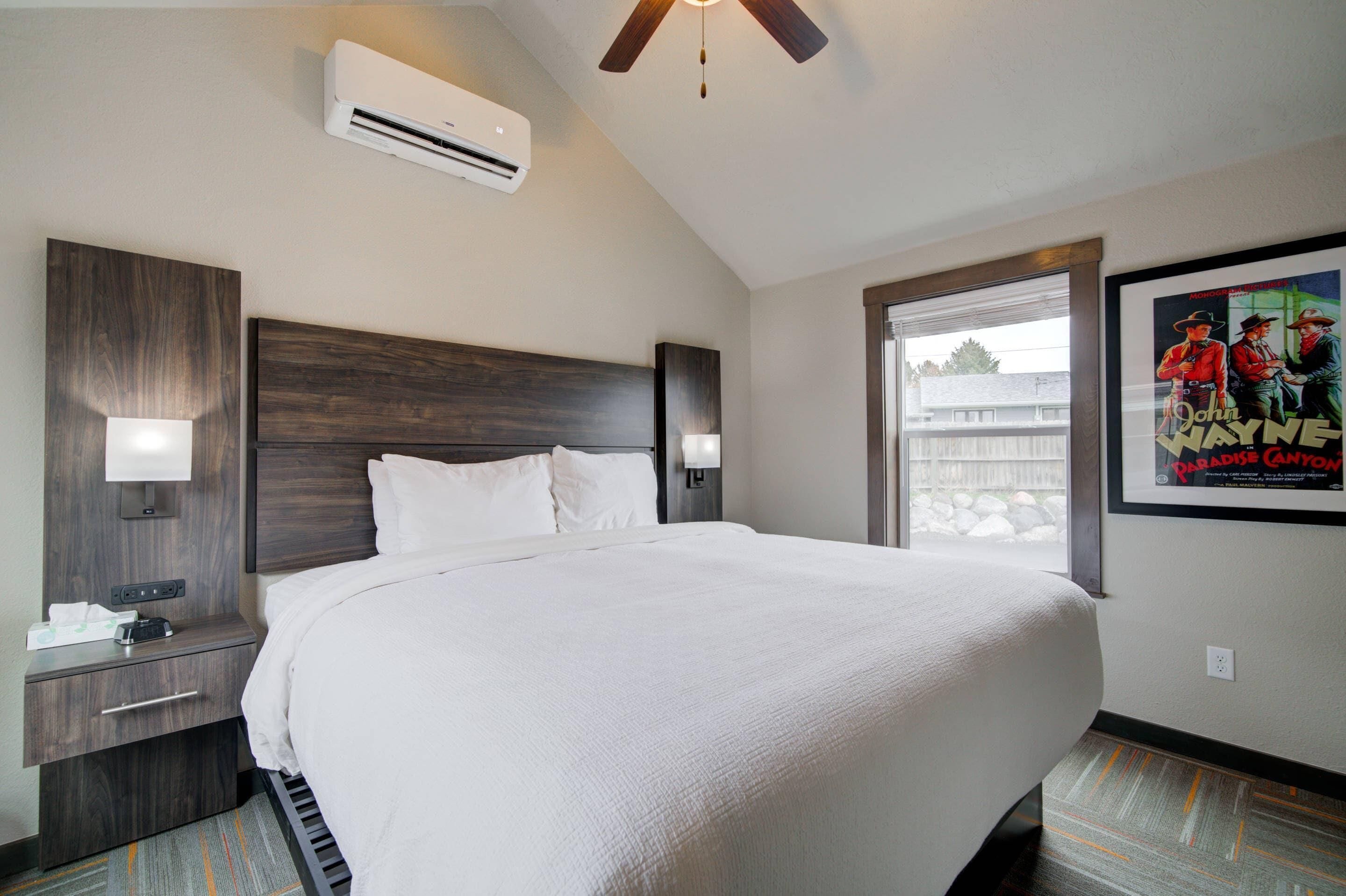 A hotel room with a large bed, a nightstand with a lamp, an air conditioning unit, and a poster on the wall.