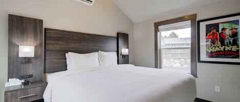 A hotel room with a large bed, a nightstand with a lamp, an air conditioning unit, and a poster on the wall.