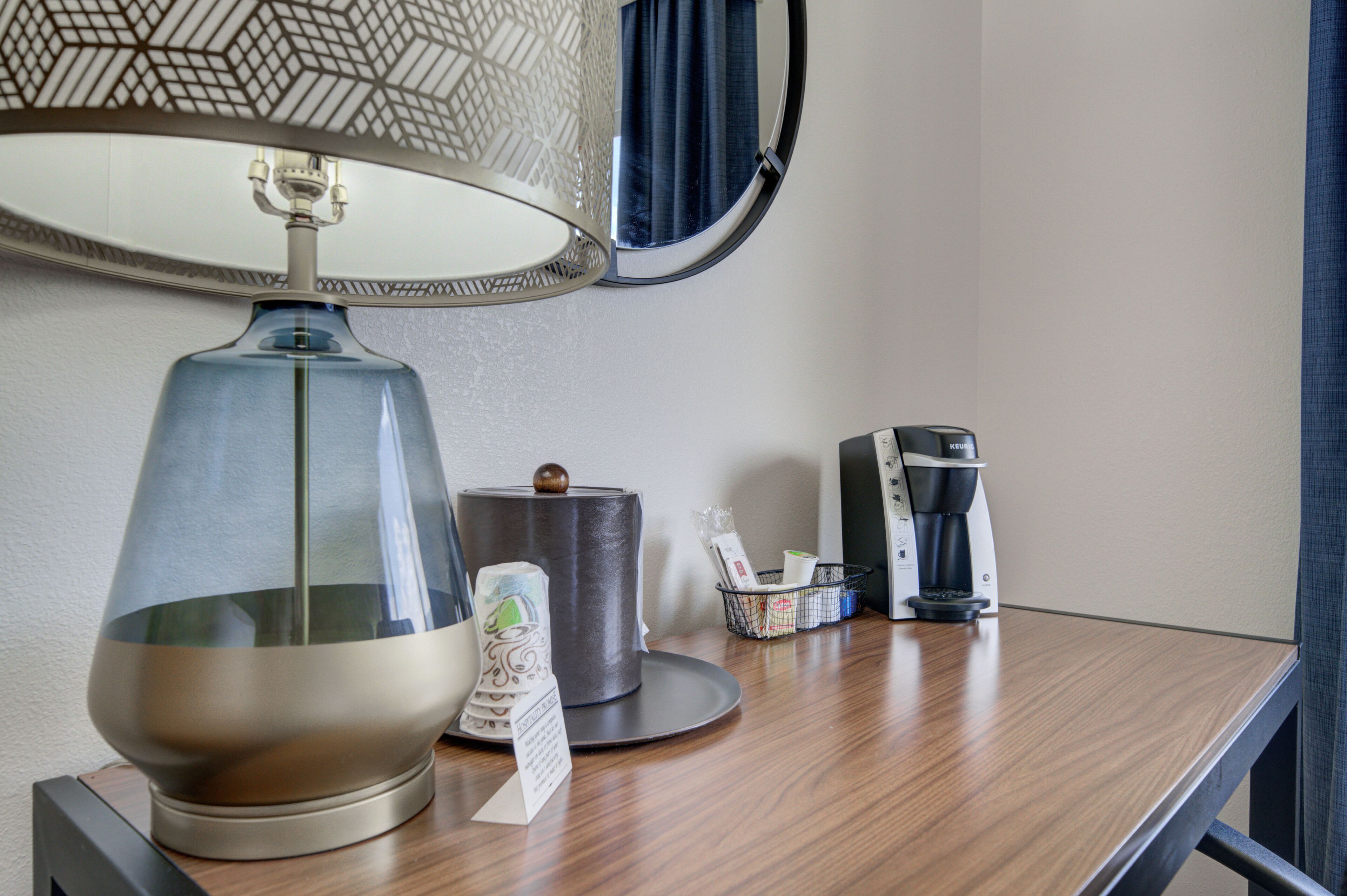 A hotel room with a wooden desk, a lamp, a coffee maker, a waste basket, and a mirror.