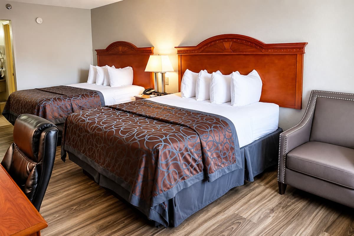 A hotel room with two beds, a wooden headboard, a desk, a leather chair, and a lamp.