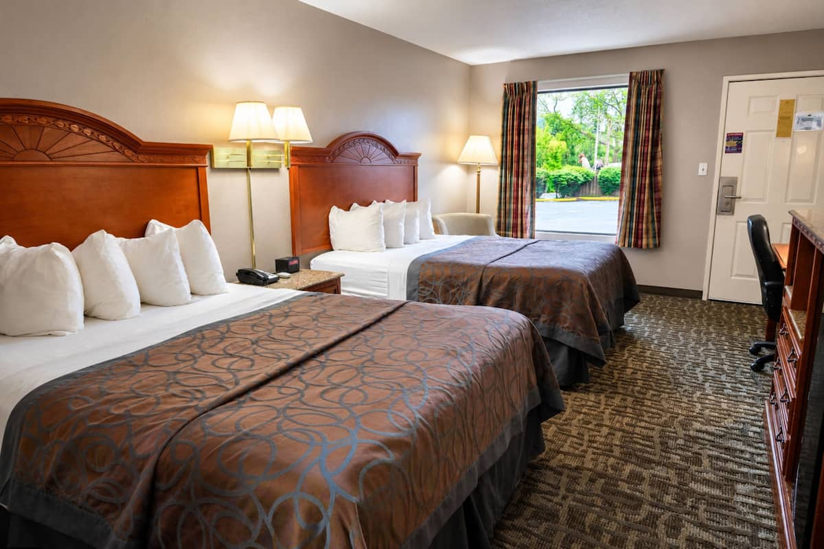 A hotel room with two beds, a desk, a chair, a lamp, and a view of the outdoors.