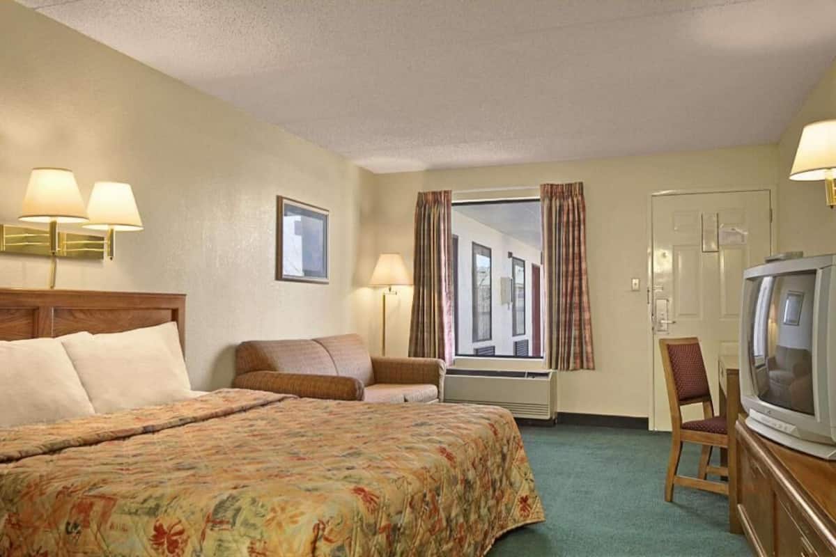 A hotel room with a bed, a television, a chair, a window with curtains, and wall-mounted lamps.