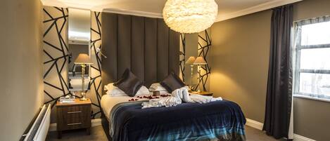 A hotel room with a large bed, two bedside lamps, a modern chandelier, and a view of the city through a window.