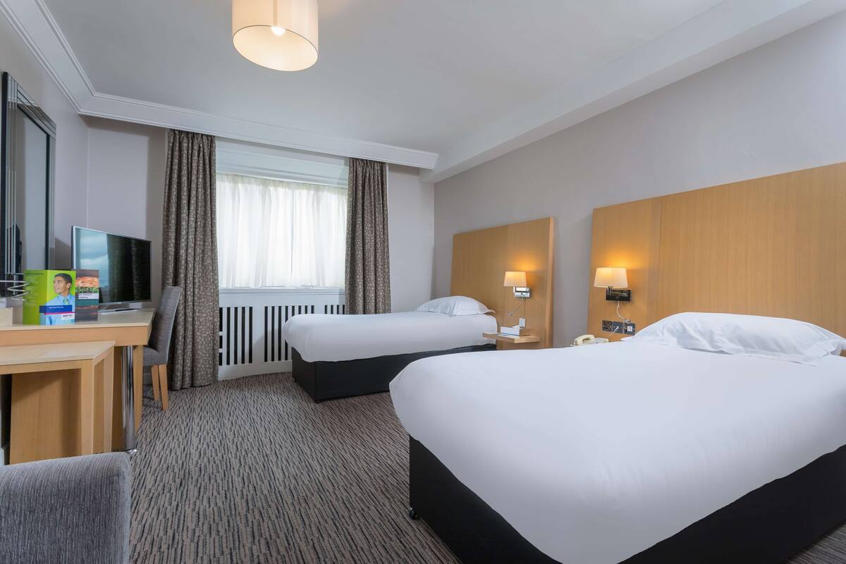 A hotel room with two beds, a desk with a TV, a lamp, and a window with curtains.