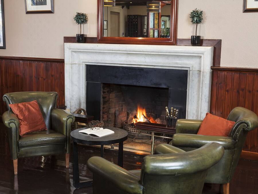 A cozy seating area with leather armchairs and a small table, a fireplace with a fire, and a mirror above it.