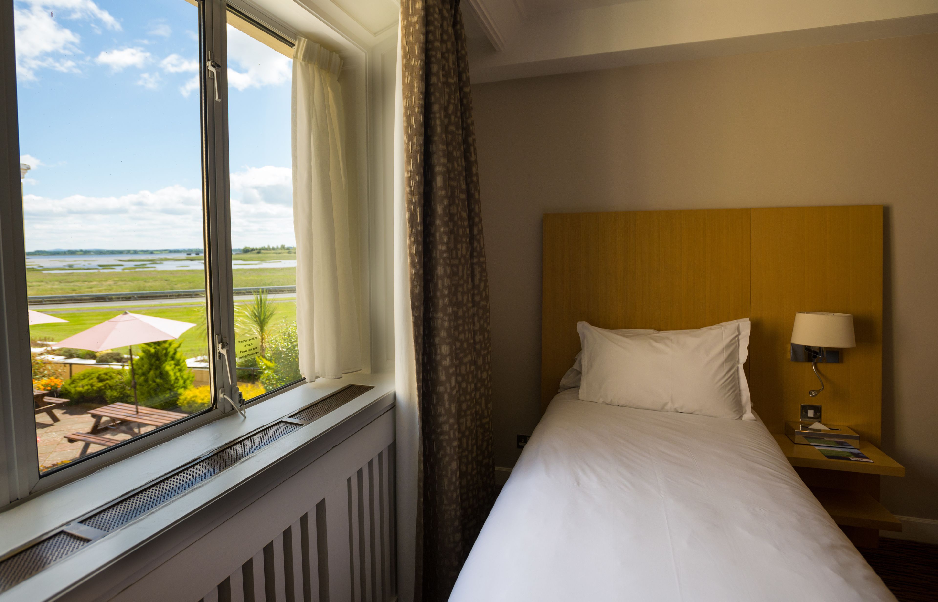 A hotel room with a large window, a bed, a nightstand with a lamp, and a view of an outdoor area with umbrellas and a picnic table.