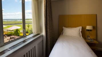 A hotel room with a large window, a bed, a nightstand with a lamp, and a view of an outdoor area with umbrellas and a picnic table.