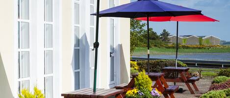 Een terras buiten met houten tafels en banken, omringd door plantenpotten en parasols.
