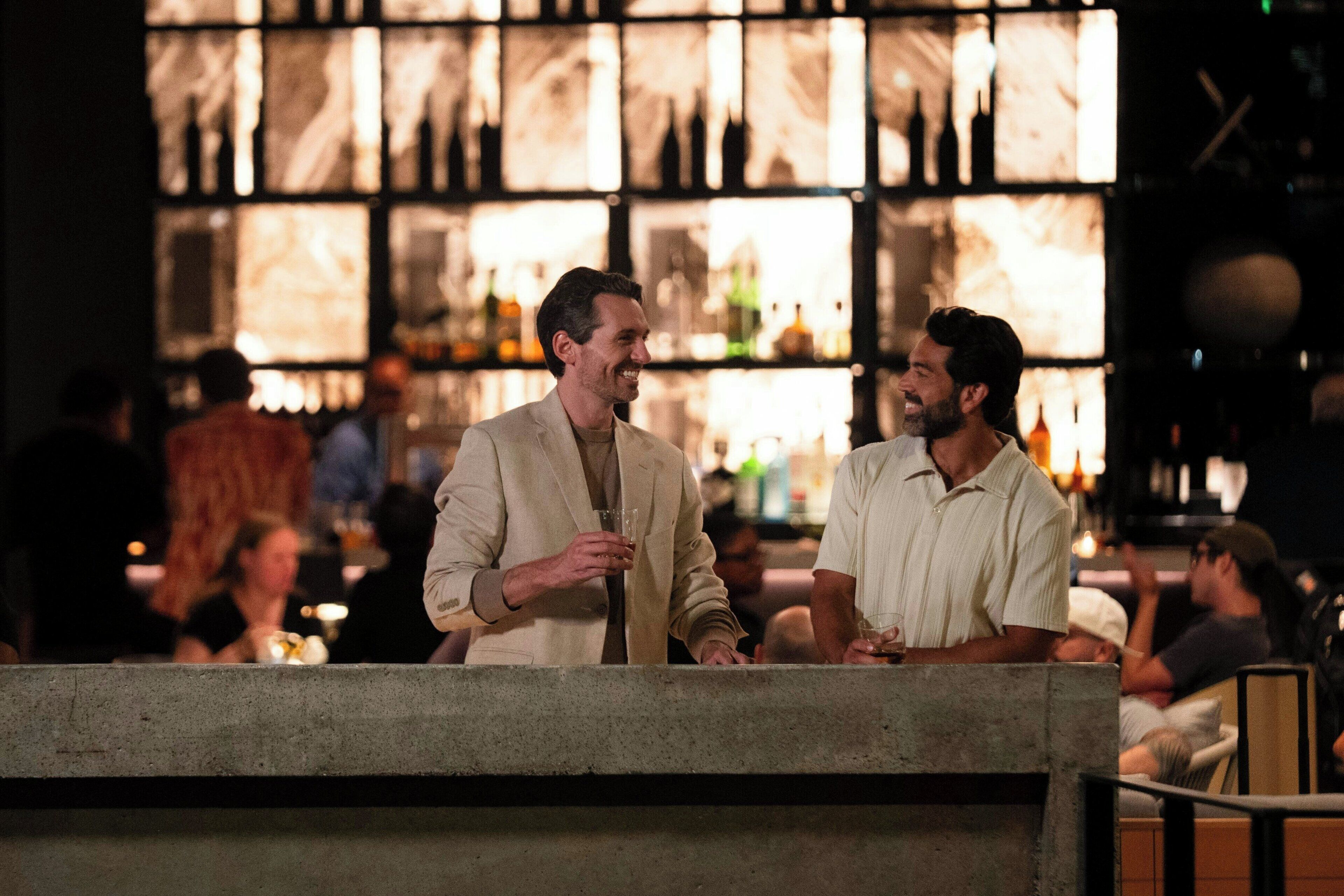 Two men conversing at a bar counter with a backdrop of shelves displaying bottles.