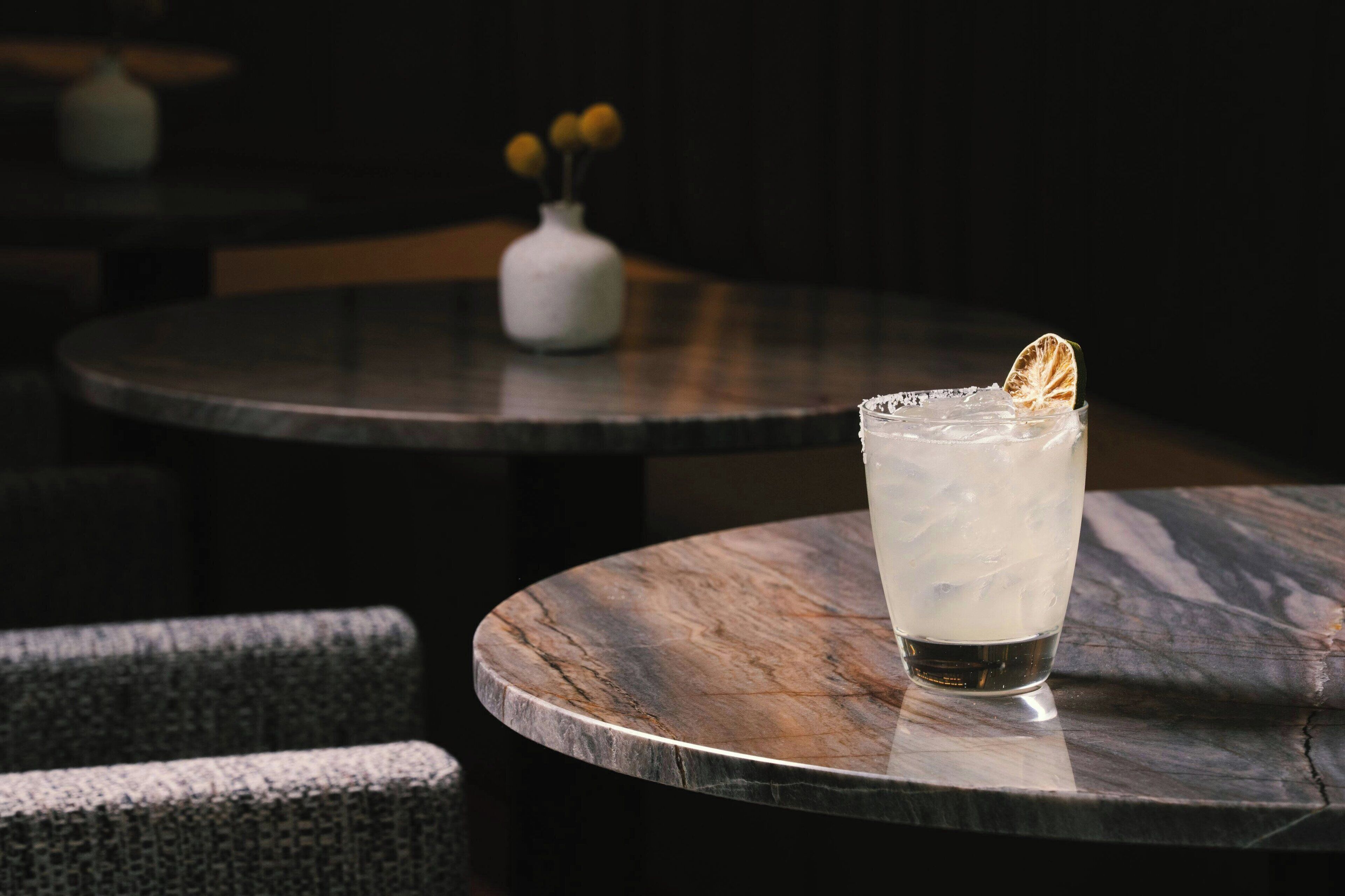 A glass of iced drink on a marble table with a blurred background of a white vase and a chair.
