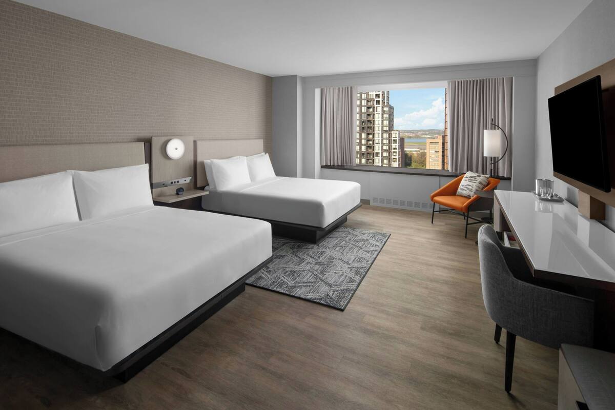 A hotel room with two beds, a desk, a chair, a TV, and a window with a city view.