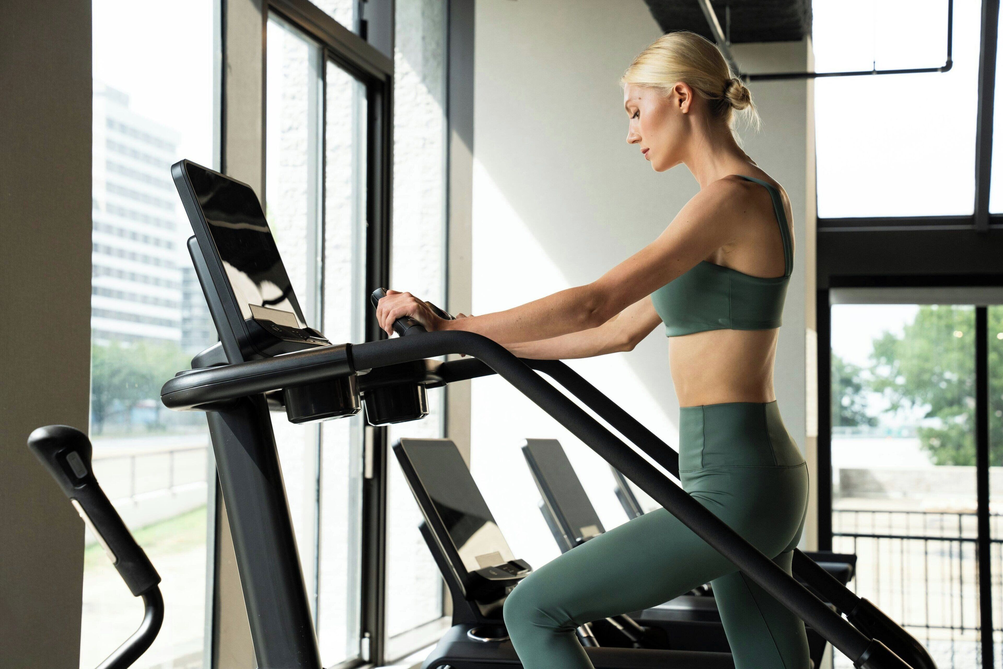 A person using a treadmill in a gym with large windows and a view of buildings outside.