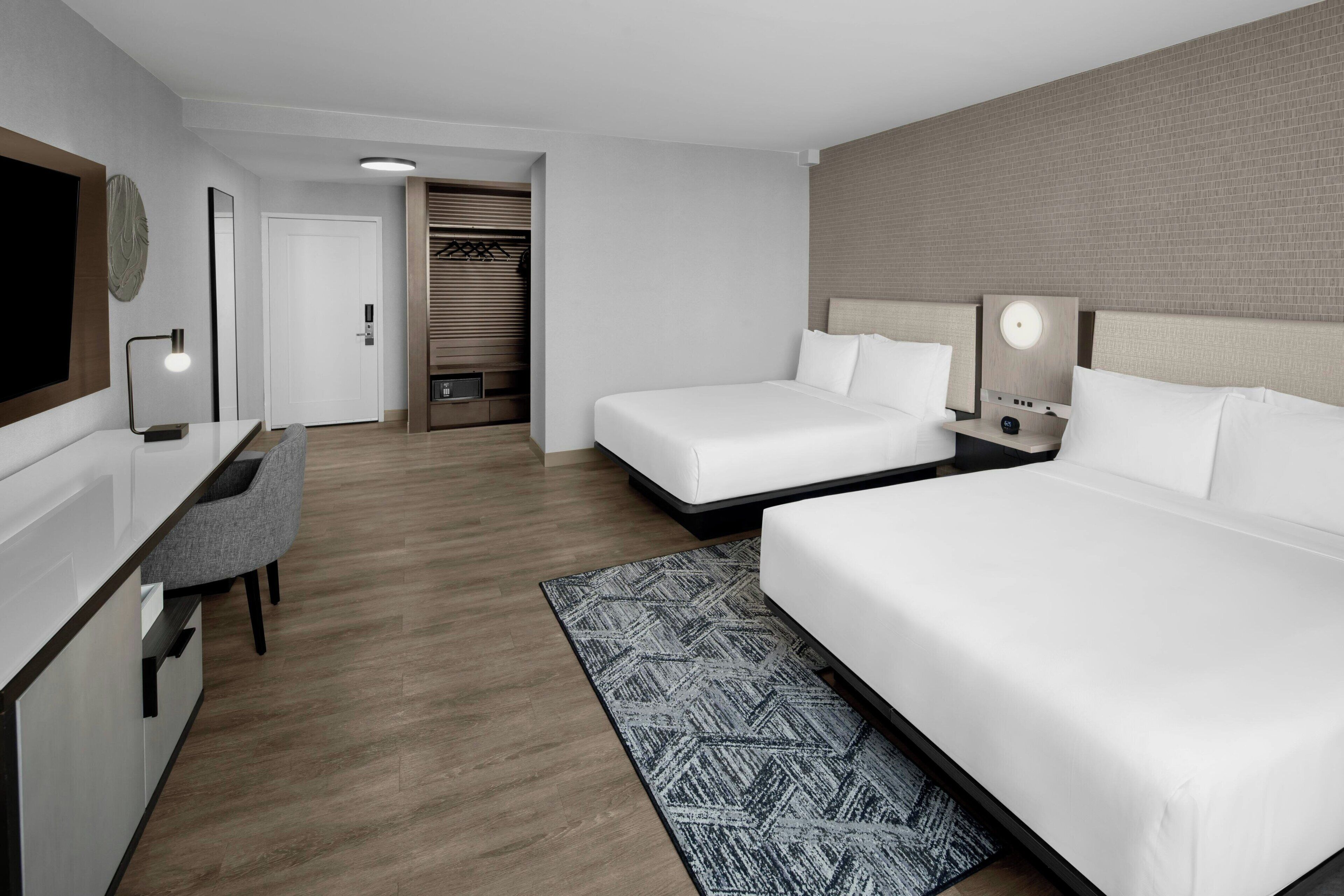 A modern hotel room with two beds, a desk, a chair, and a wardrobe.