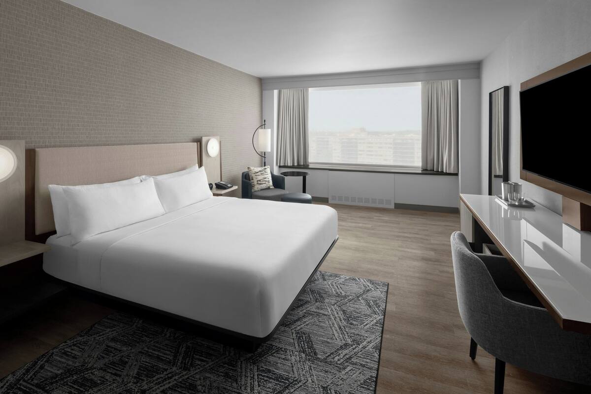 A modern hotel room with a large bed, a desk, a chair, a TV, and a window with curtains.