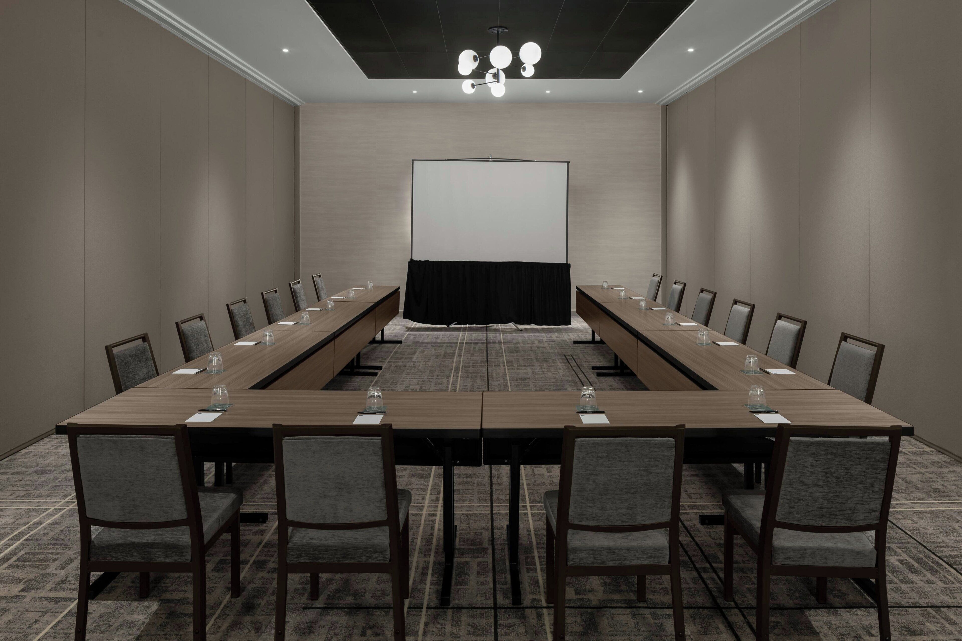 Uma sala de conferências com uma mesa comprida, cadeiras, uma tela de projetor e um lustre.