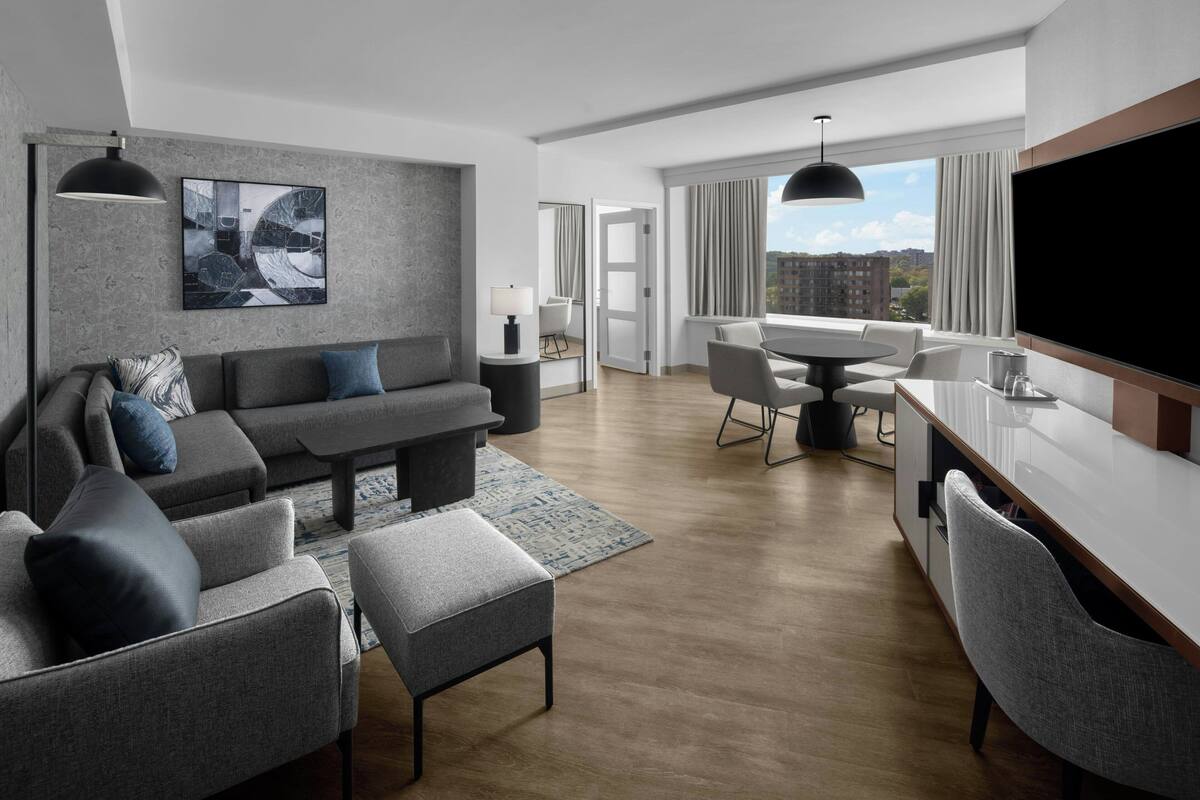 A modern living room with a sofa, armchairs, a coffee table, and a dining area with a table and chairs. There is a TV mounted on the wall and a large window with curtains.