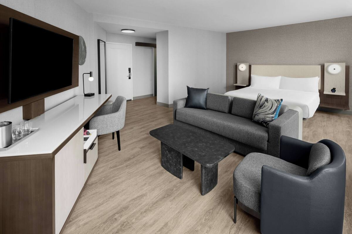 A modern hotel room with a flat-screen TV, a sofa, a bed with two pillows, a coffee table, and a desk with a lamp.