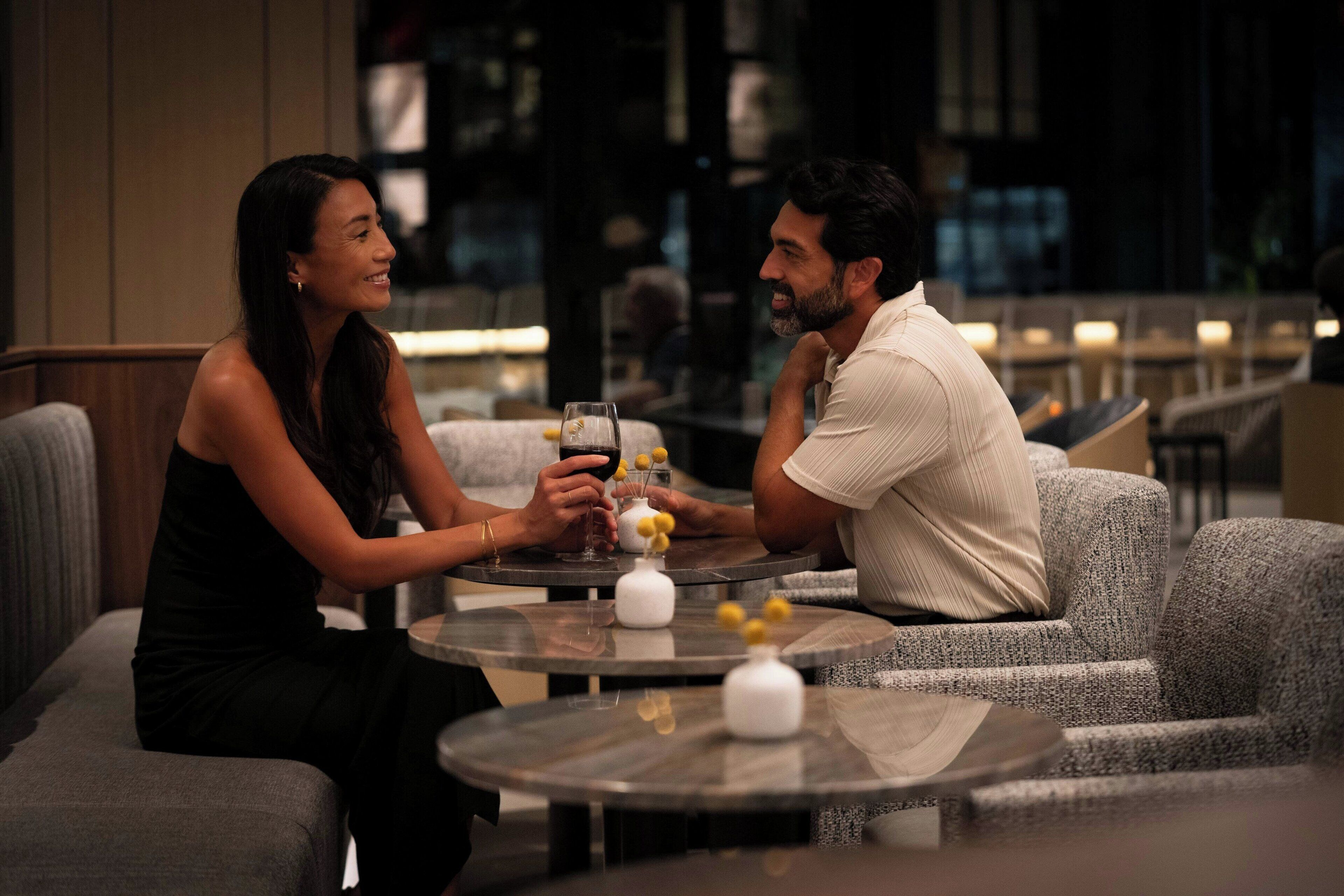 A couple seated at a round table in a dimly lit indoor setting, possibly a restaurant or lounge area, with a modern interior design and comfortable seating.