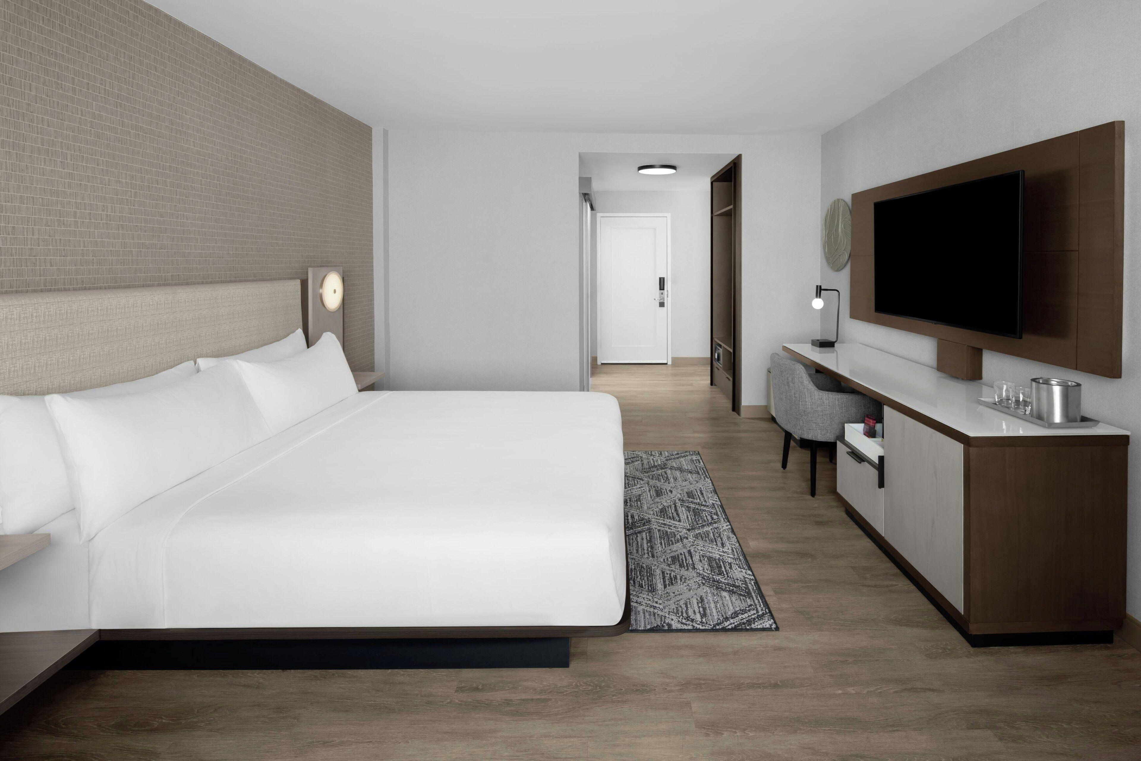 Una habitación de hotel moderna con una cama grande, un televisor montado en la pared, un escritorio pequeño y un vestidor.