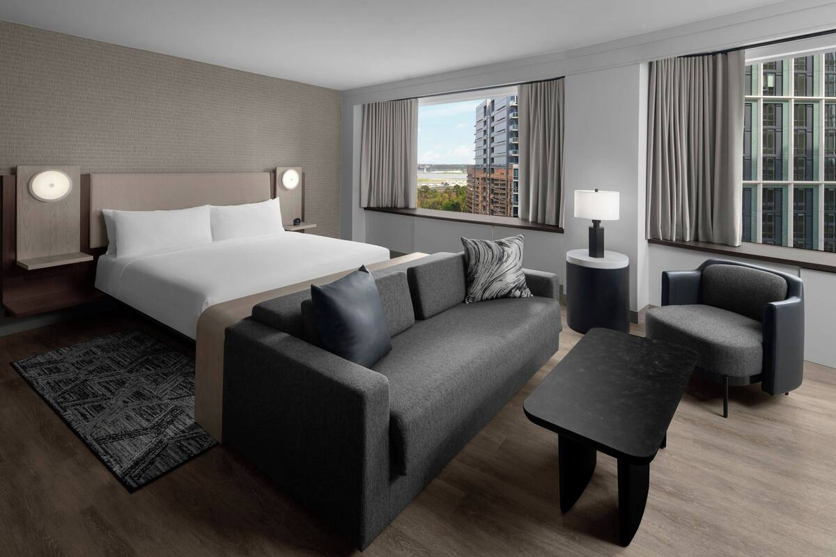 A modern hotel room with a large bed, a sofa, a coffee table, and a chair. There are two bedside tables with lamps, a window with curtains, and a view of buildings outside.