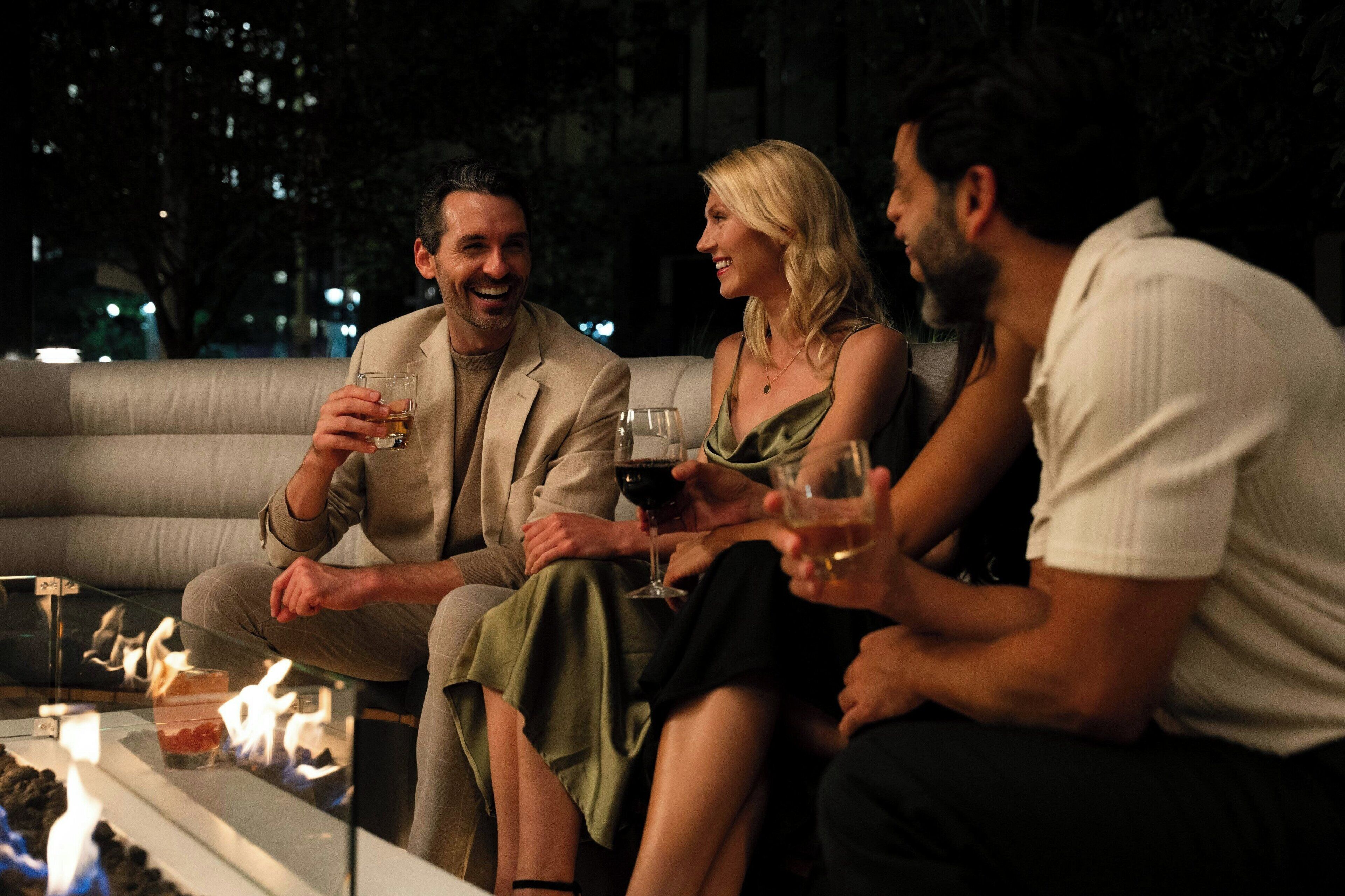 A group of people seated on a couch around a table with a fire, holding drinks.