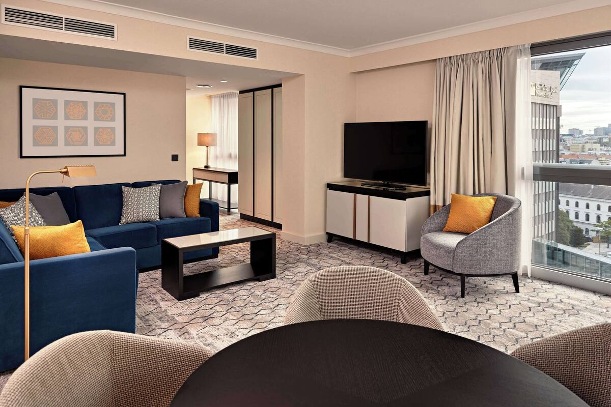 A modern hotel room with a sofa, armchair, TV, and a city view.