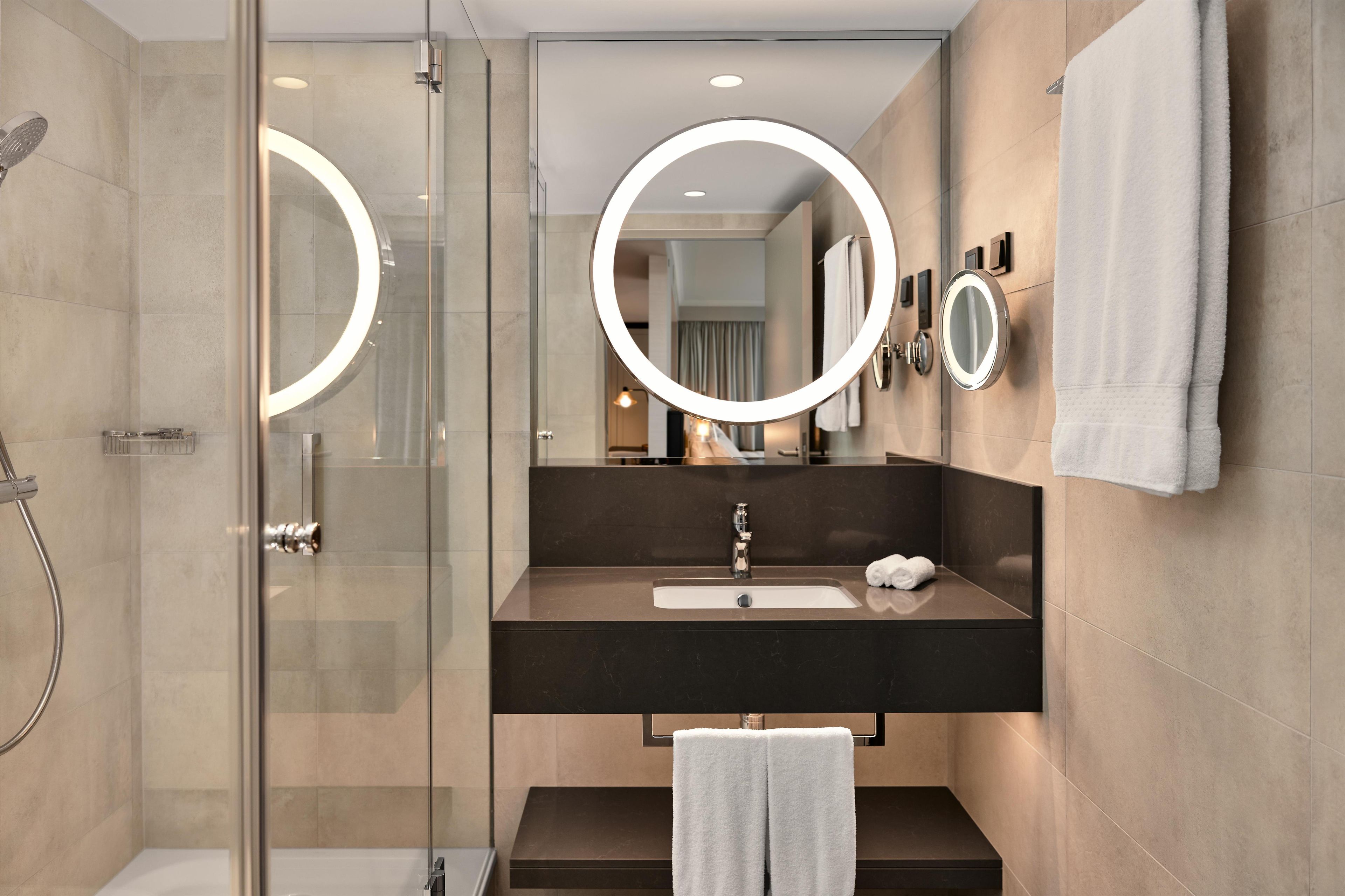 Een moderne badkamer met een douchecabine van glas, een wastafel met donker aanrechtblad en twee wandspiegels met verlichte omlijstingen.