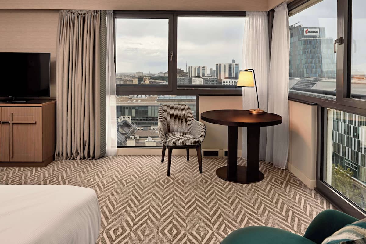 A hotel room with a large window, a television, a patterned carpet, a chair, a small table, and a bed.