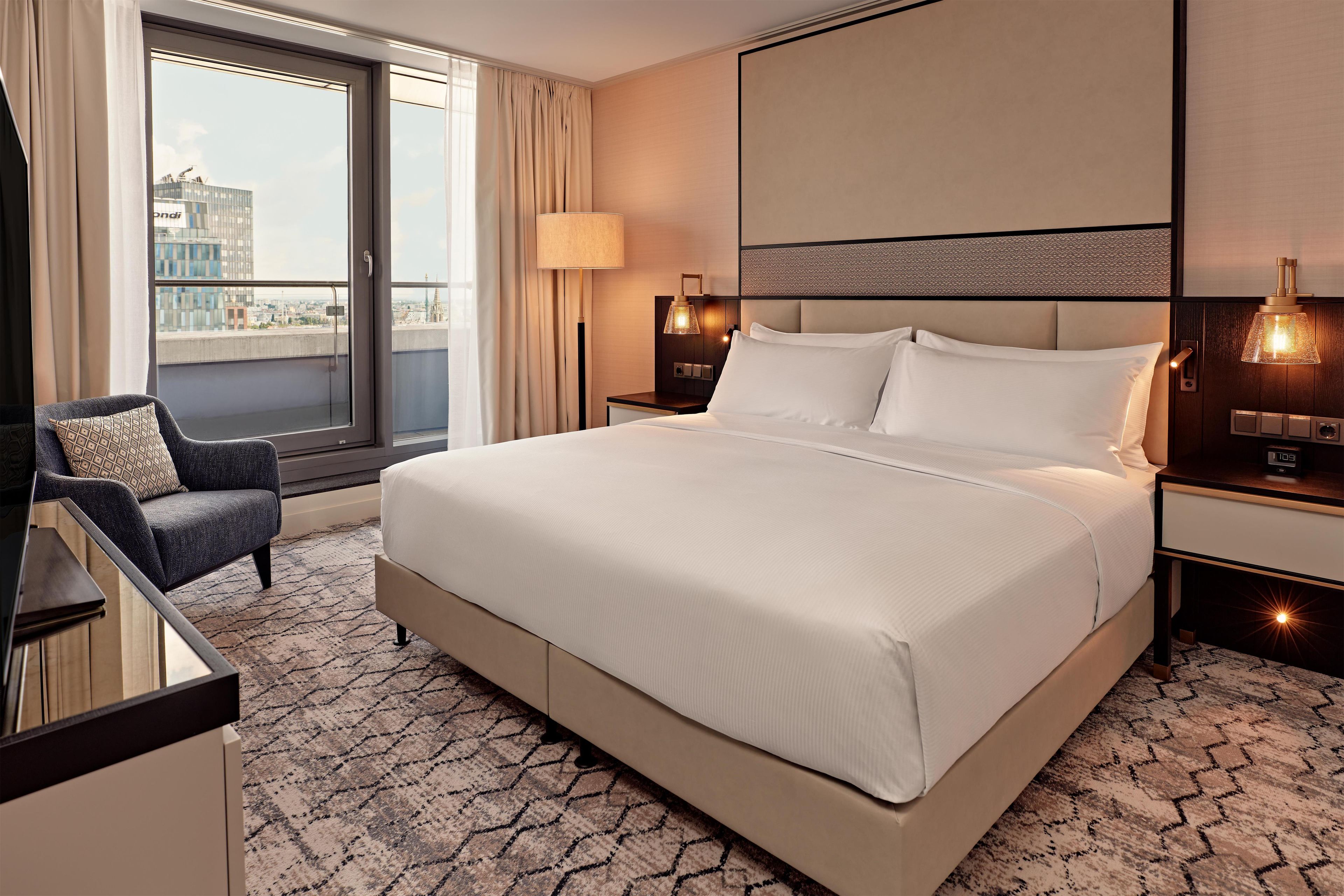 Ein Hotelzimmer mit einem großen Bett, einem Sessel, einem Schreibtisch und Blick auf die Stadt.