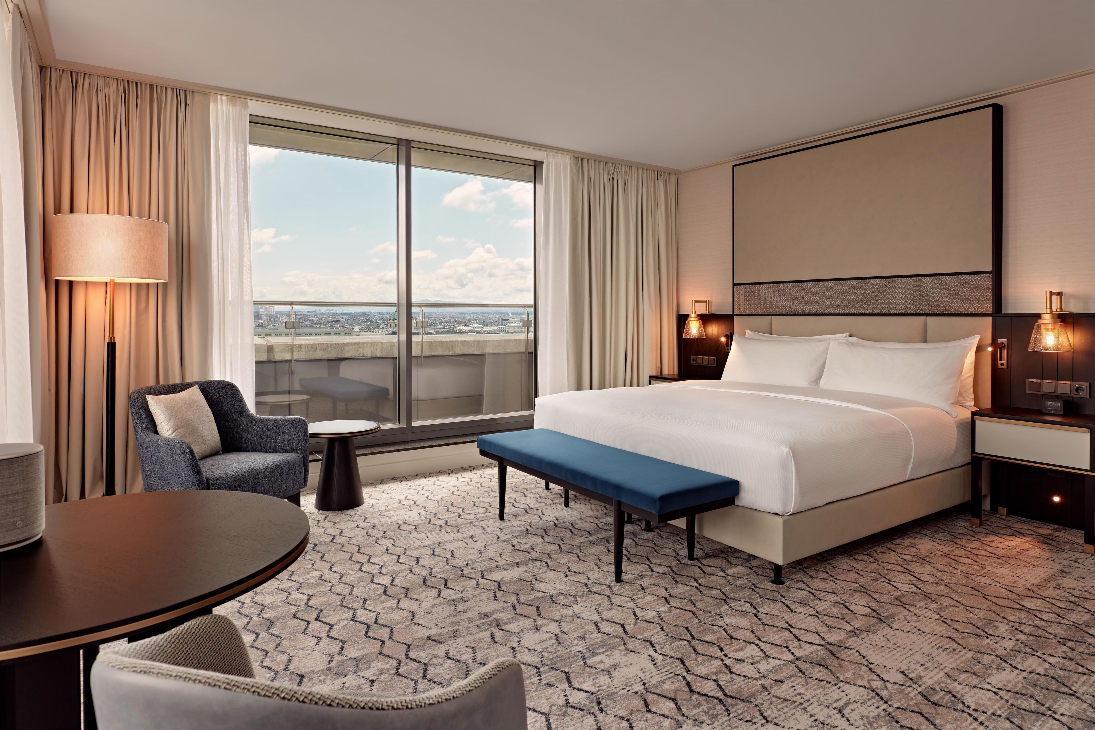 A hotel room with a large bed, a desk, a chair, a bench, and a view of the city.