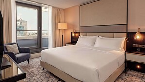 A hotel room with a large bed, a desk, a chair, a lamp, and a view of the city through the window.