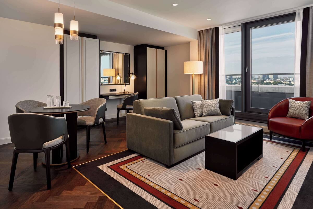 A modern hotel room with a sofa, armchair, dining table, and a large window offering a city view.