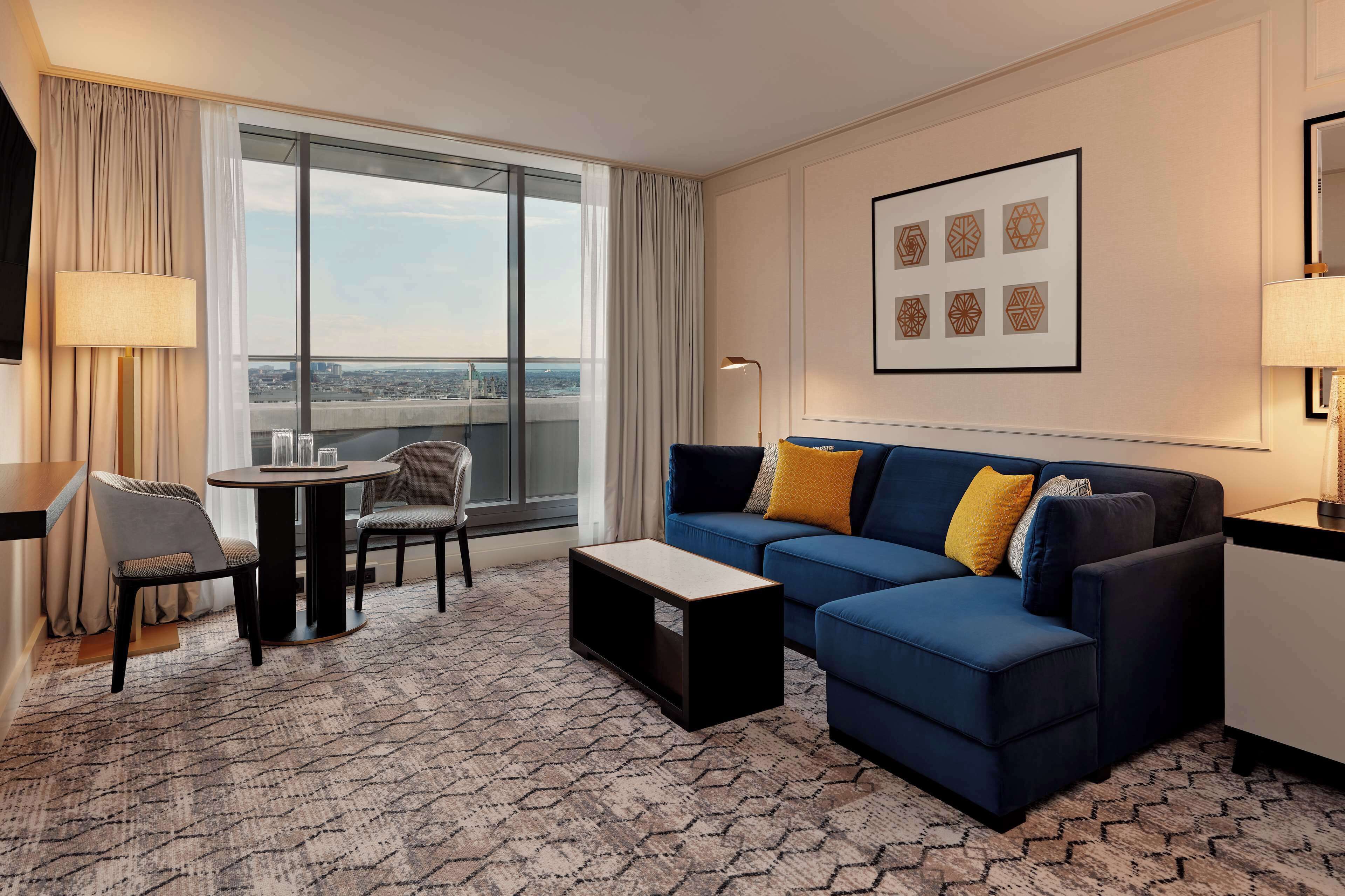 Een moderne hotelkamer met een groot raam, een blauwe bank met gele kussens, een klein rond tafeltje en een kamerbreed tapijt met een patroon.