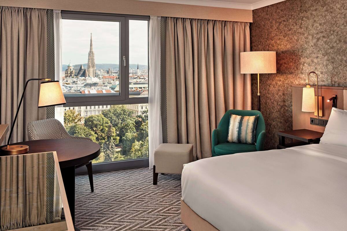 A hotel room with a large bed, a desk, a chair, and a view of a cityscape through a large window.