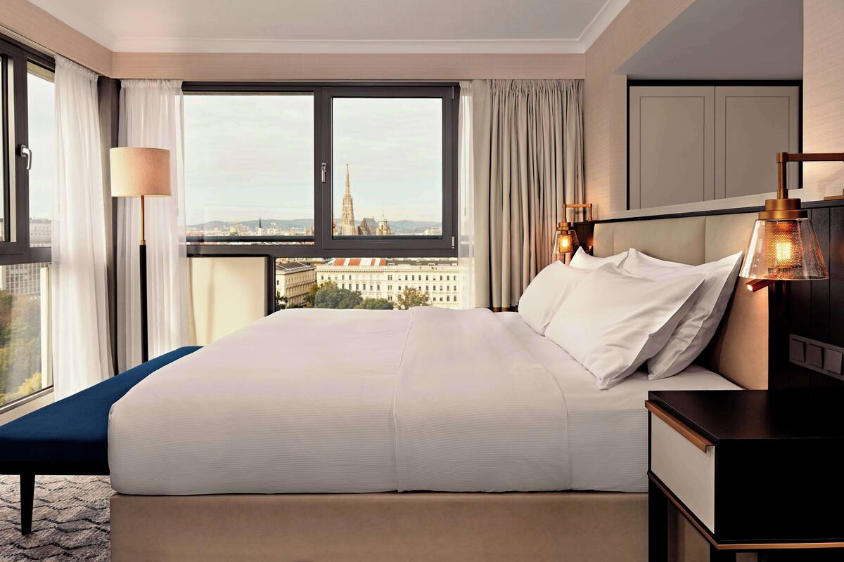 A hotel room with a large bed, a view of a cityscape, a bedside table, and a lamp.