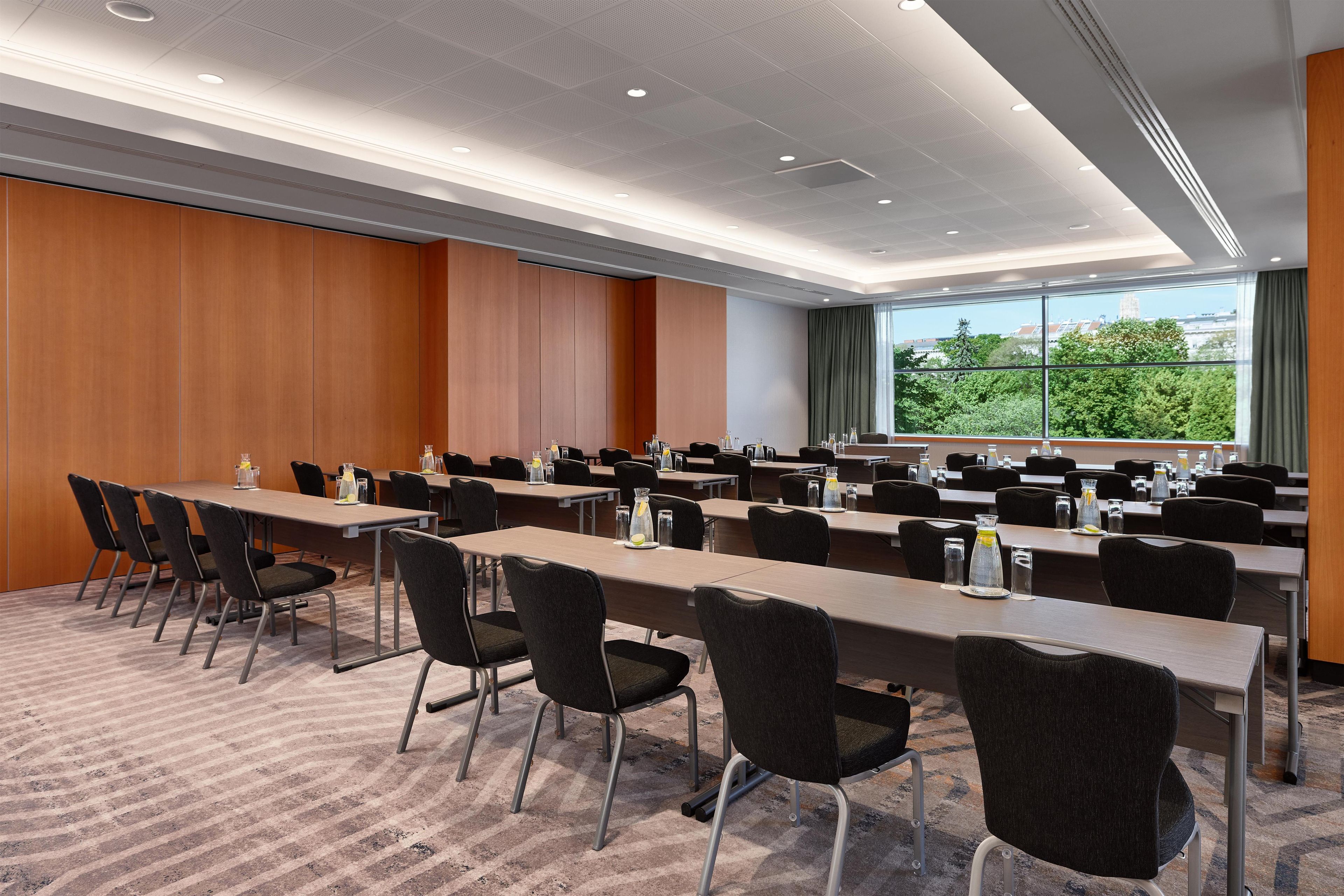 Une salle de conférence dotée de plusieurs tables et chaises, de grandes fenêtres offrant une vue sur la verdure et d’un plafond avec des luminaires encastrés.