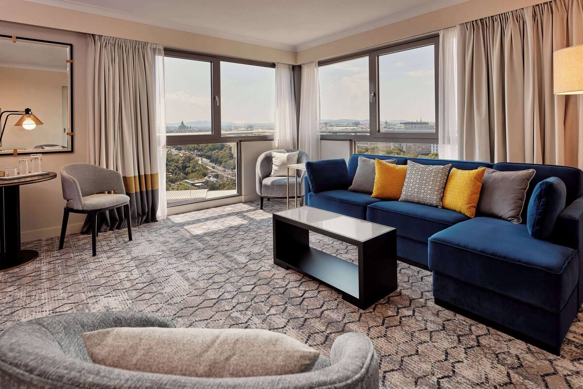A modern hotel room with a large window, a blue sofa, a grey armchair, a small table, and a patterned carpet.