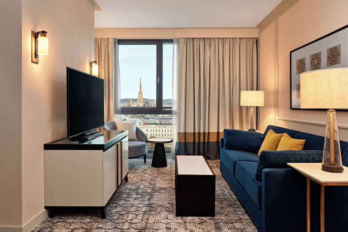 A hotel room with a large window, a flat-screen TV, a sofa, a coffee table, and a lamp.