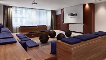 Una habitación de hotel moderna con una pantalla grande que muestra 'Hilton Vennna Park', un mostrador de recepción de madera y una zona de estar con cojines azules y pelotas de ejercicio.