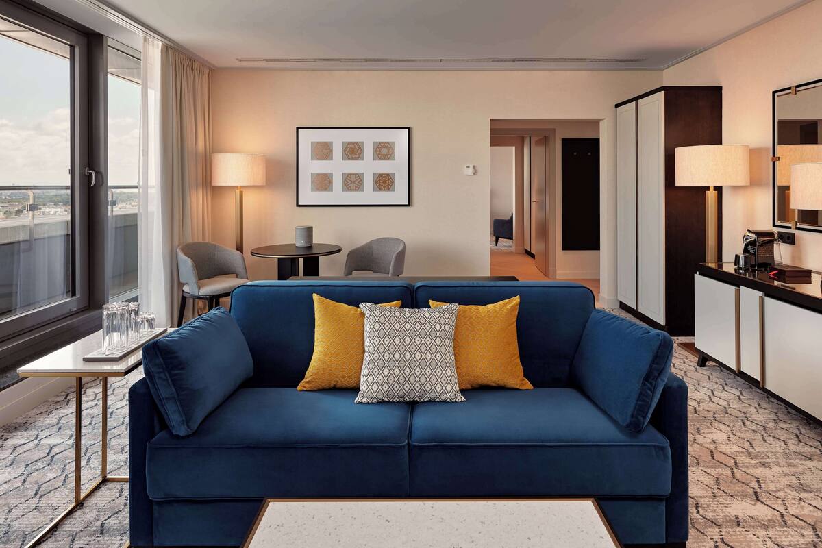 A modern hotel room with a blue sofa, a small table with glasses, a lamp, and a framed picture on the wall.