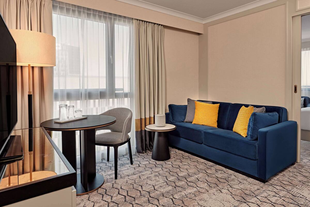 A modern hotel room with a blue sofa, a round table, a chair, and a lamp.