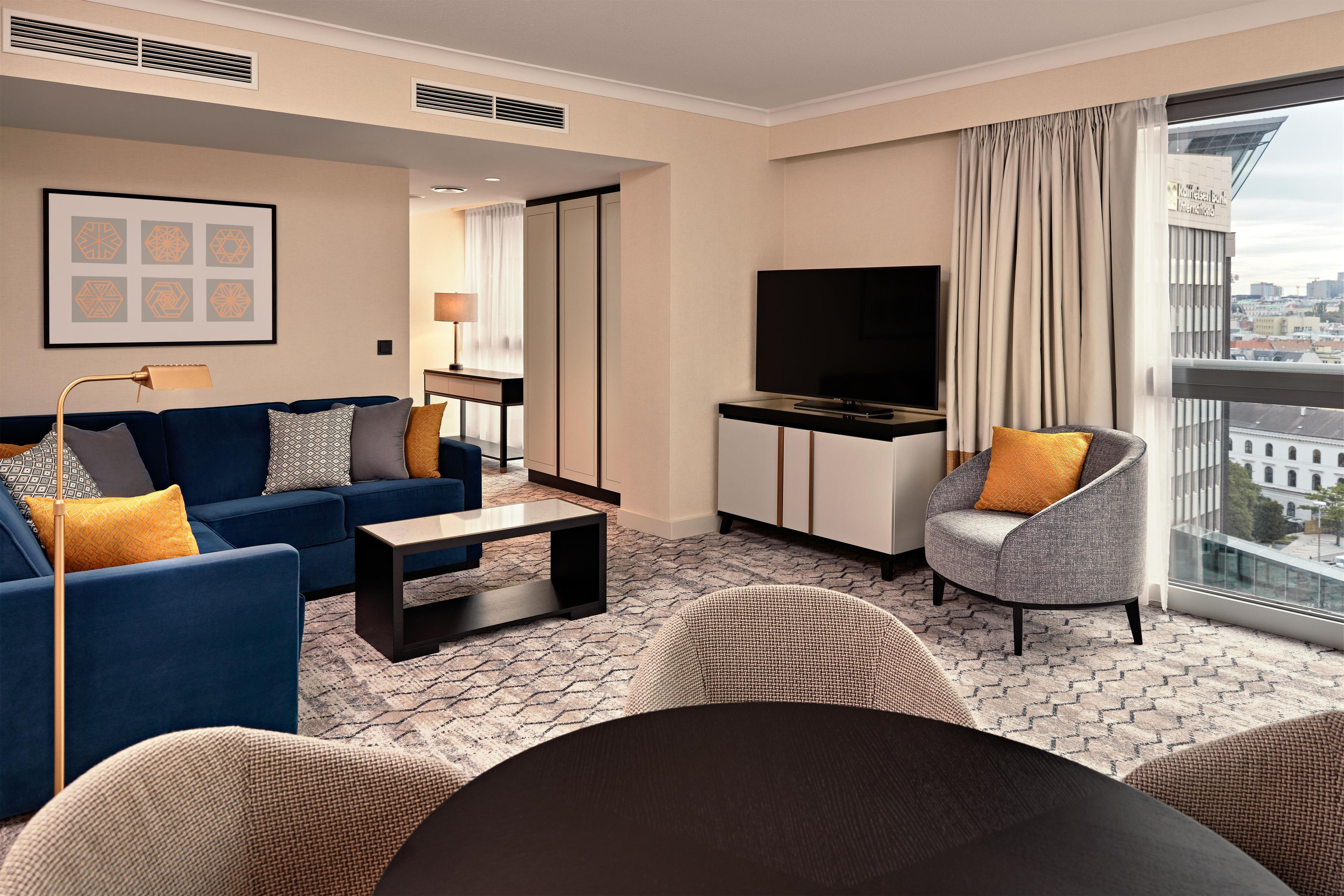 Ein modernes Hotelzimmer mit Sofa, Sessel, Fernseher und Blick auf die Stadt.