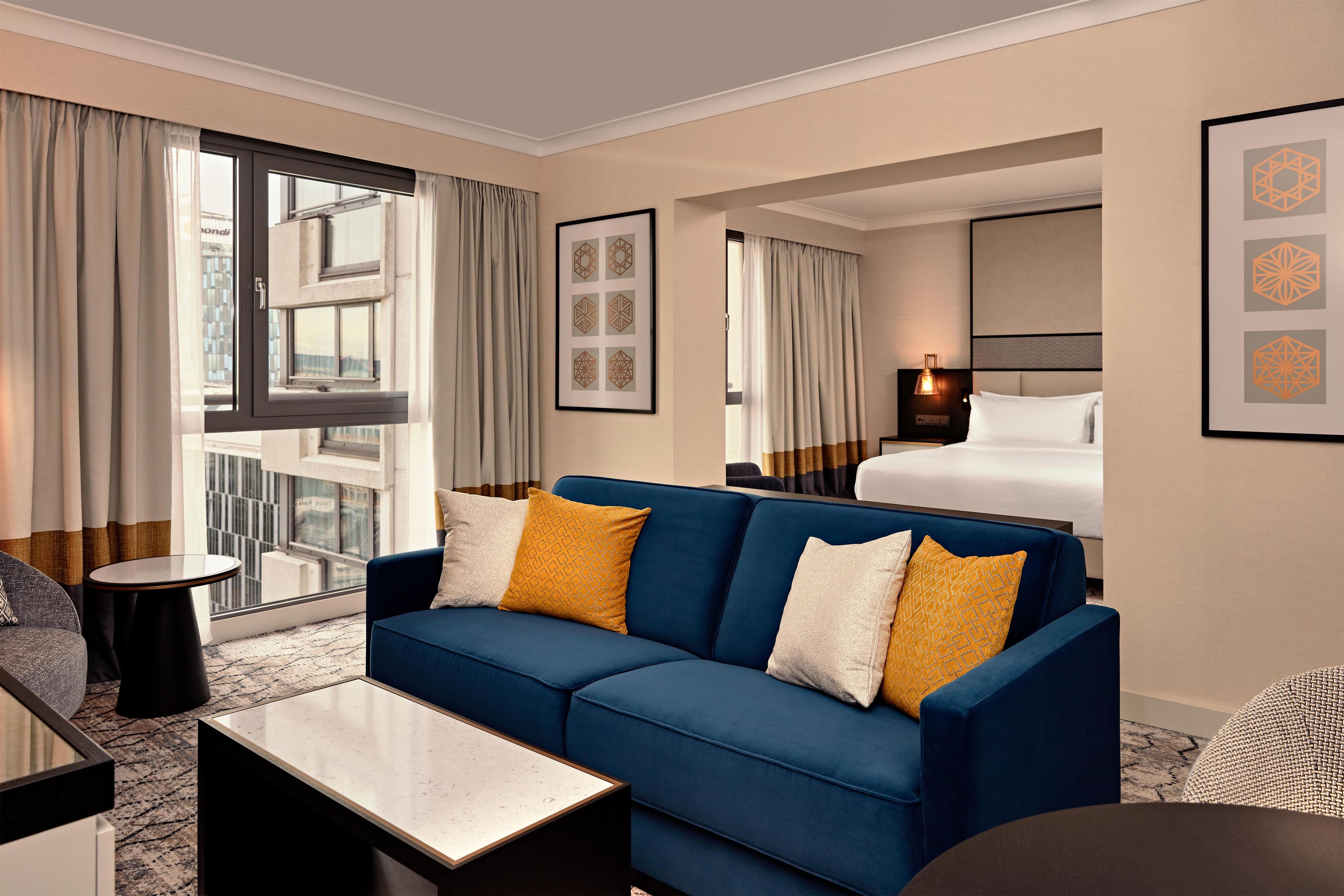 Ein Hotelzimmer mit blauer Couch, einem kleinen Tisch, einem Bett und Blick auf die Stadt durch große Fenster.