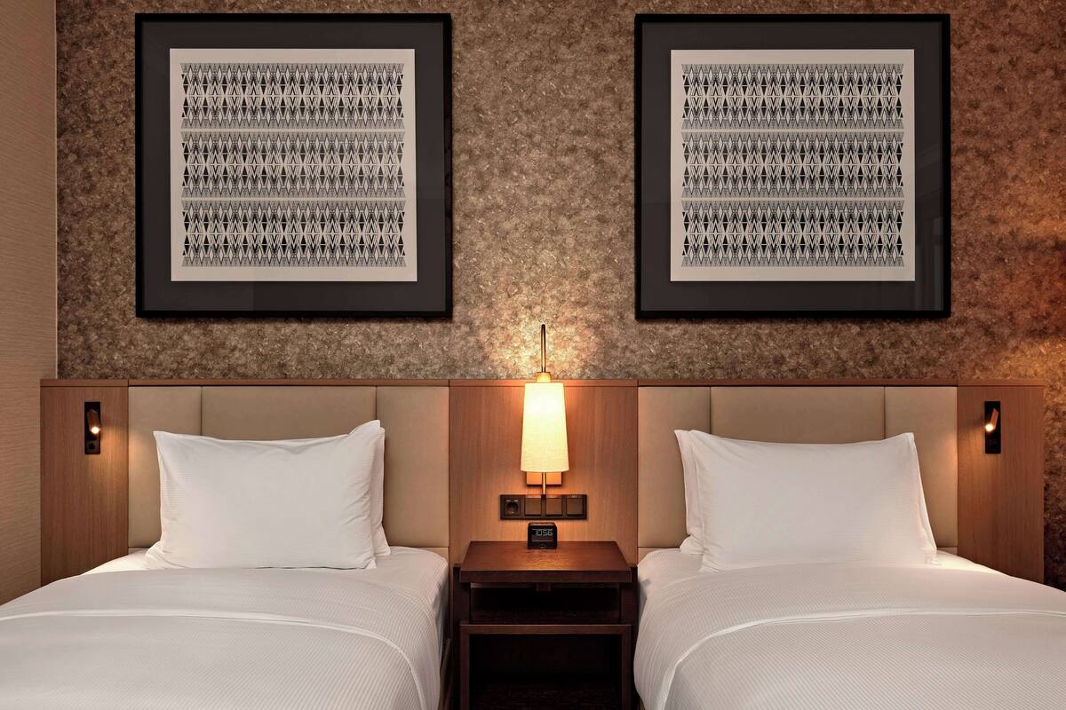 A hotel room with two beds, a nightstand with a lamp, and two framed geometric wall art pieces.