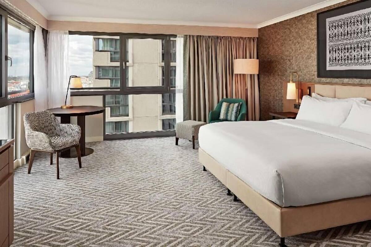 A hotel room with a large bed, a desk, a chair, a small table, a lamp, a view of buildings through a window, and a patterned carpet.